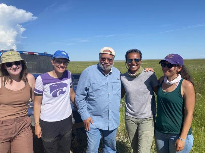 Five adults pose for a photo in a lush field-like area with a big blue sky above and a vehicle or structure slightly obscured in the background on left. In center is Lonnie Bunch III, the Secretary of the Smithsonian, in denim with cap.