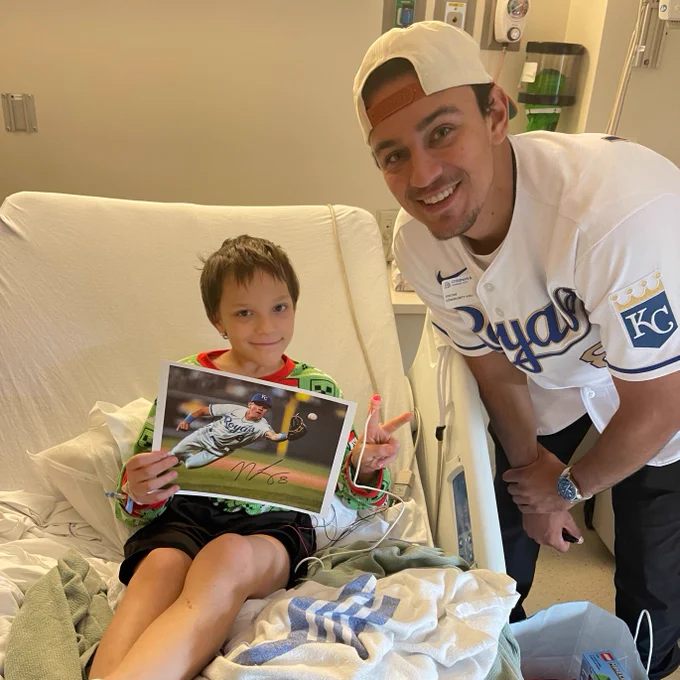 Riley is sitting in his hospital bed holding up an autographed photo of Nicky and making a peace sign. Nicky is standing next to him smiling.