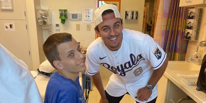Kyler is sitting in his wheelchair smiling and Nicky is standing next to him smiling.