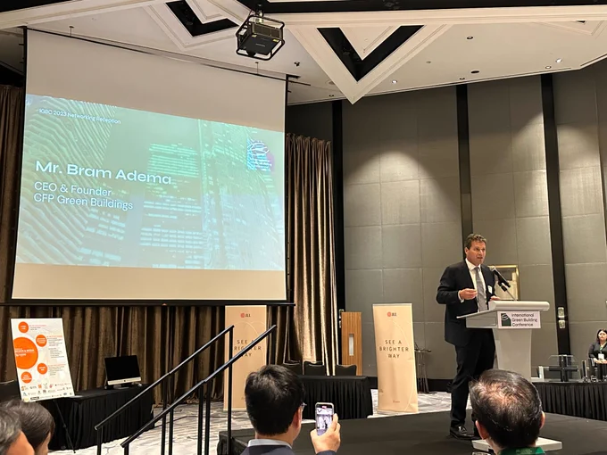 Snippet from the conference, there is a man in black suit standing at the podium talking to the audience. Behind him is a screen which displays, "Mr. Bram Adema, CCEO & Founder, CFP Green Buildings". 