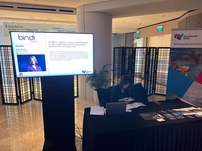 Snippet from the conference; there is table with a black cloth behind which a woman is seated and can be seen working on her phone. On the table, there many pamphlets placed, along with papers and a laptop. On left of the table, there is a screen which shows BindiMaps profile. There is bindiMaps Logo on top, beneath is an image of Anna Wright (our co-founder) and next to it is some information about BindiMaps and Anna. On right of the table, there is a pullup banner of Investment NSW
