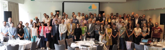 A large group of people stands in a room in front of a screen. The screen displays the EOSC-Life logo.