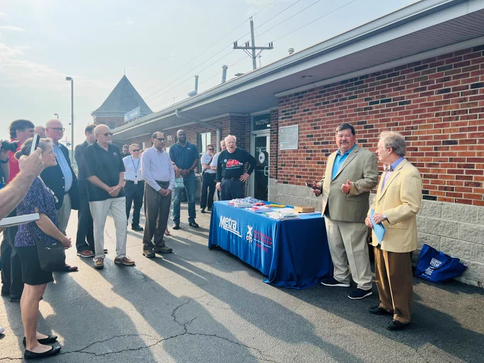 Mayor Rod Craig and male guest providing remarks outside in front of a red brick train station with a blue metra table on the side while onlookers listen.