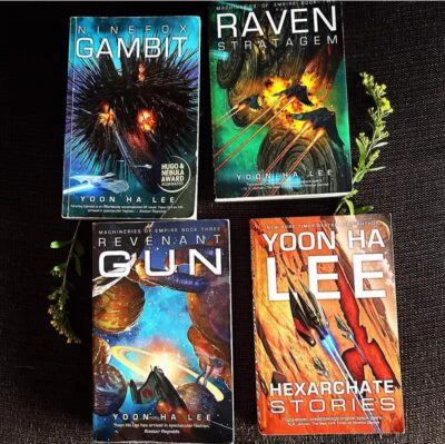 Ninefox Gambit, The Raven Stratagem, Revenant Gun and Hexarchate Stories by Yoon Ha Lee, lying face-up on a black surface surrounded by some flowers.