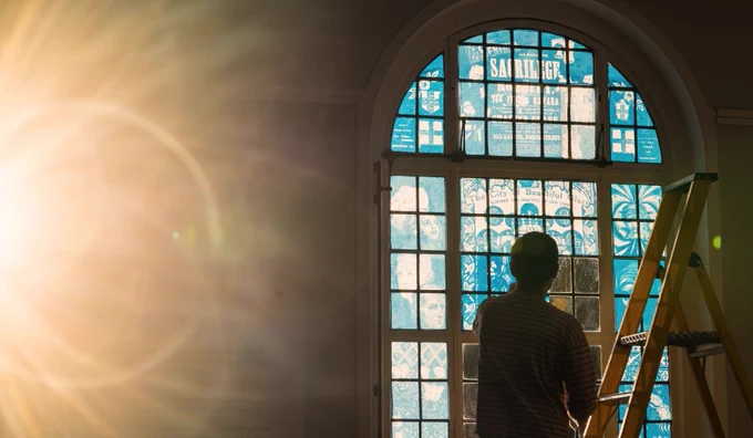 Photograph of a silhouette of a person at the top of a step ladder in front of an arched window. The window is covered with transparent cyanotype images of transparencies held in our archive