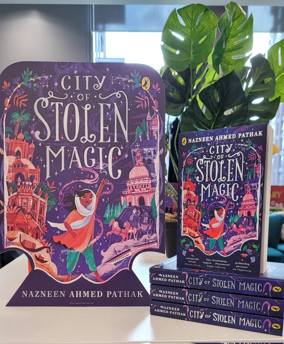 Copies of the book City of Stolen Magic are stacked on the right, with a large cardboard cutout of the purple and silver book cover on the left 