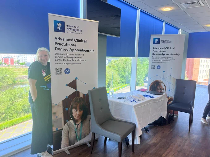 A member of the University of Nottingham's ACP Team smiles at the camera and stands with the university's ACP Degree Apprenticeship banners in front of a window in a conference room at the Leicester King Power Stadium. 

The banner reads' Advanced Clinical Practitioner Degree Apprenticeship
Designed to meet advanced clinical skills requirements across the healthcare industry.' and shows a mental health nursing student taking a patients blood pressure.  There is a table and chairs next to the banner with information leaflets and pens on the table. 