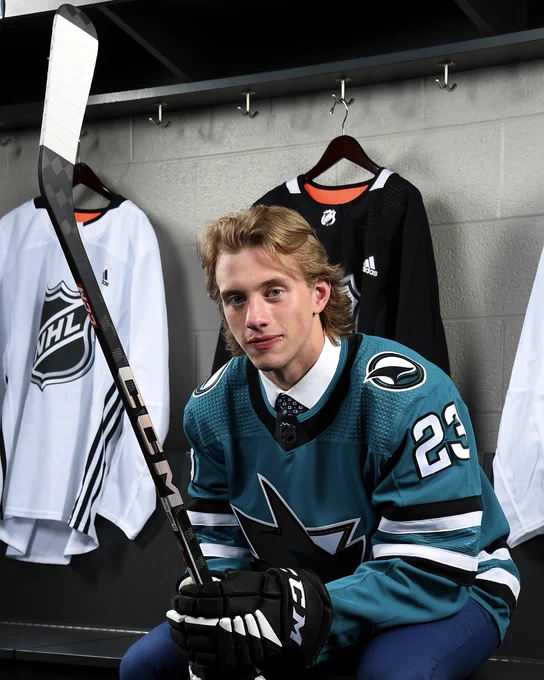 Picture of Quentin Musty looking up and grinning, wearing a Sharks jersey and holding a hockey stick. There are 3 NHL hockey jerseys on the wall behind him.