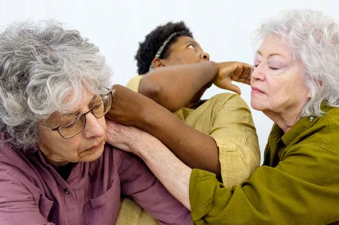 Three elderly women share a joyful moment, laughing and embracing each other warmly in casual clothing.