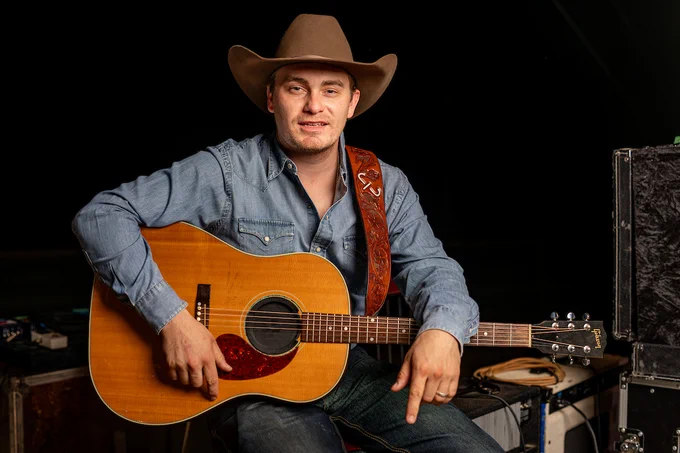 Man wearing a cowboy hat and holding a guitar smiles and poses for a picture. 