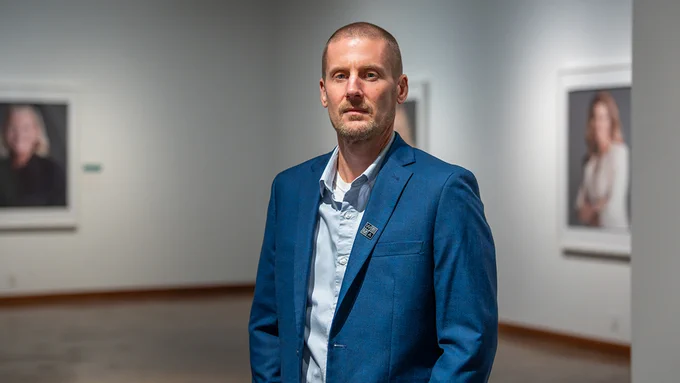 Matthew Bailey wearing a blue blazer stands confidently in an art gallery with portraits on the wall in the background.