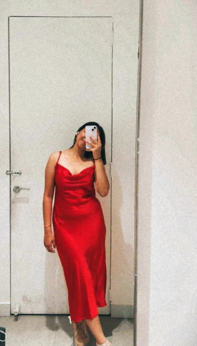 A woman stands in front of a full-length mirror in a clean, minimalist room, taking a mirror selfie that captures her from head to toe. She is wearing a sleek red satin dress with thin straps and a softly draped neckline, the fabric flowing smoothly over her silhouette. The dress falls to mid-calf, creating an elegant, timeless look. Her face is partially hidden by her phone, adding a sense of mystery and intimacy to the image.  

The neutral background and simple door behind her keep the focus on the rich red color of the dress and the calm, self-assured energy she radiates. The overall mood feels sensual, poised, and quietly powerful, with an emphasis on feminine confidence rather than overt display.