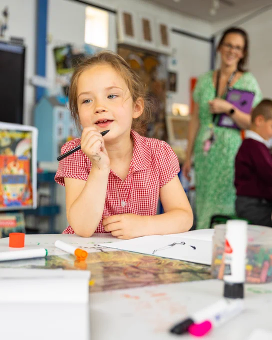 Young girl in a red gingham dress sits at a table with art supplies, deep in thought during a classroom activity.