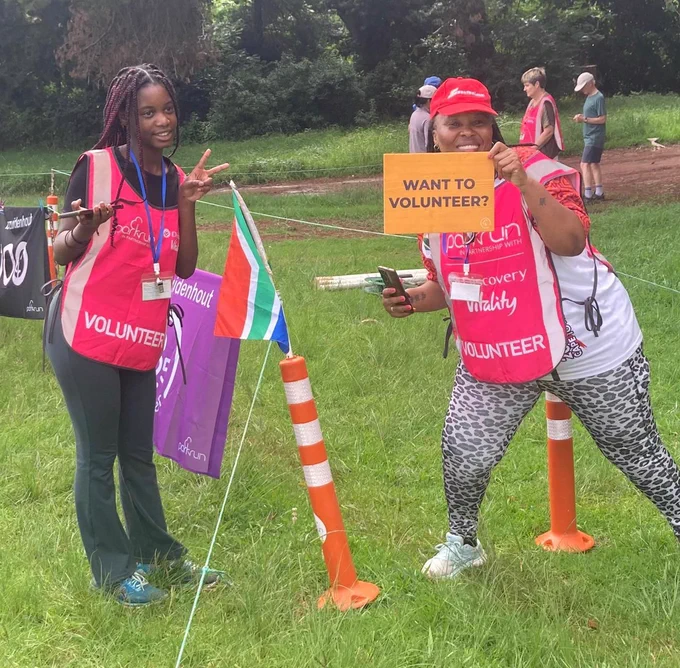 Two parkrun volunteers posing at the finish funnel excitedly, one holding a card saying 'WANT TO VOLUNTEER?' and the other gesturing a V-sign.