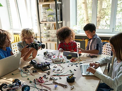 Children collaborating on a robotics project with electronic parts and laptops around a large table in a bright room.
