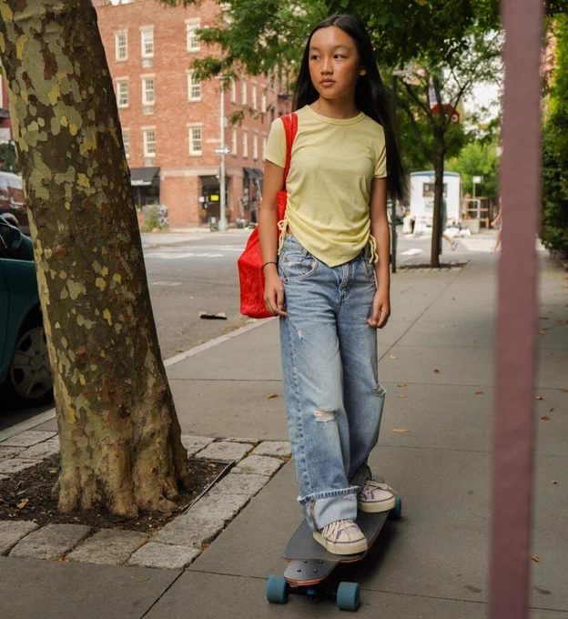 Malea Emma rides a skateboard on a city sidewalk, wearing a yellow top, ripped jeans, and a red bag over one shoulder. She looks focused amongst an urban and calm setting.