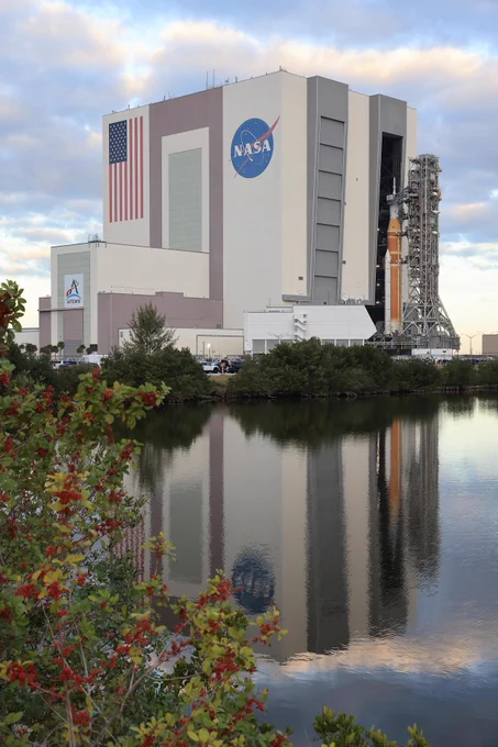 The Artemis II vehicle exits the Vehicle Assembly Building on its way to the launch pad on January 17, 2026. A reflection of the scene is seen in a body of water that makes up the foreground.