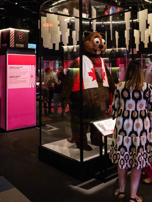 Une personne regardant une exposition présentant une grande mascotte de castor portant un t-shirt avec le drapeau canadien, exposée dans une vitrine en verre transparent dans un musée.