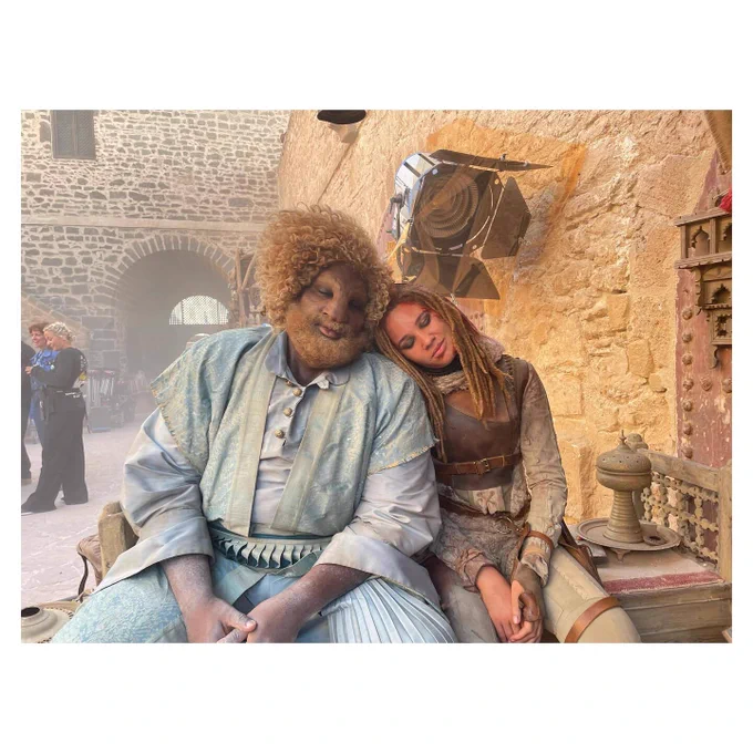 Ayoola Smart as Aviendha sits beside Hammed Animashaun as Loial on a stone bench in The Wheel of Time. Both have their eyes closed, with Aviendha resting her head gently against Loial’s shoulder. Aviendha wears worn Aiel-style clothing and long braids, while Loial, the tall Ogier, is dressed in layered pale blue and earth-toned robes with curly hair and a beard. The scene appears to be a behind-the-scenes set with stone walls and film lighting visible in the background.