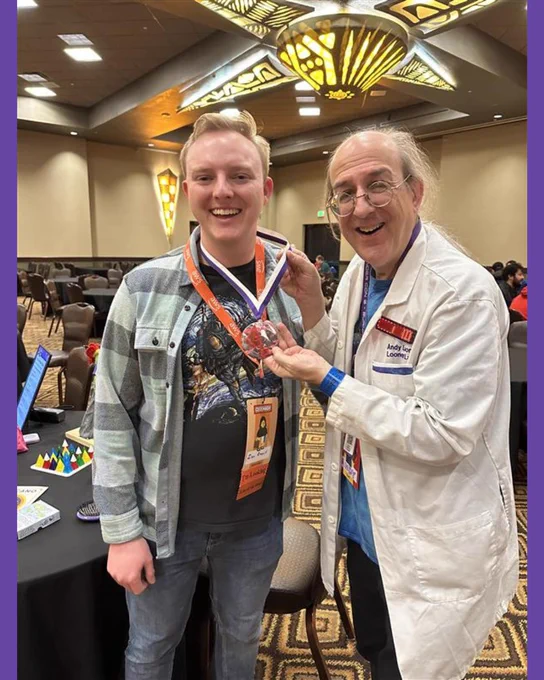 Andy in a lab coat awarding a medallion to a convention goer in a convention room with games on tables in the background