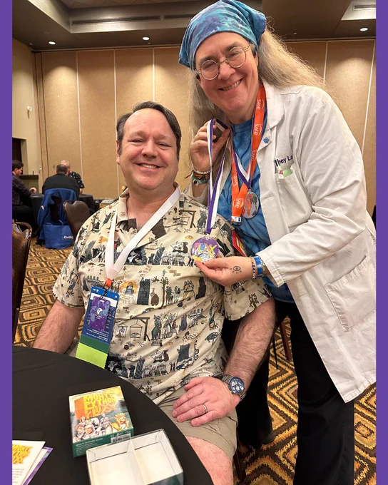 Kristin awarding a medallion to a person at a convention with Monty Python Fluxx on a table in the foreground and a convention room in the background