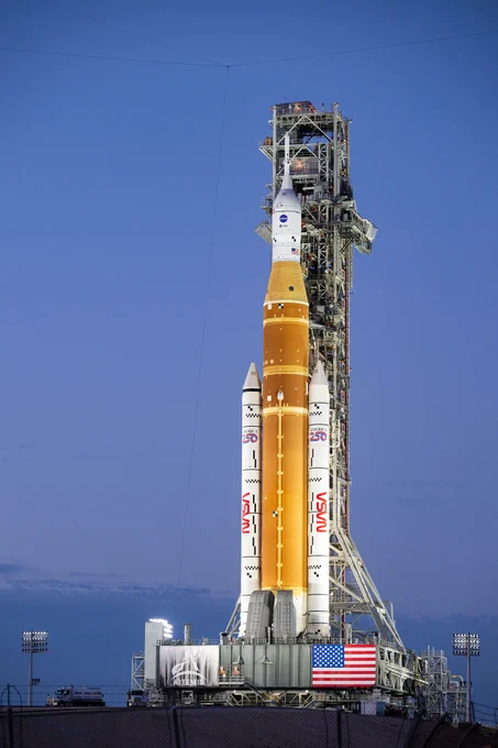 NASA’s Artemis II Space Launch System (SLS) rocket and Orion spacecraft are seen illuminated by lights at Launch Complex 39B, Saturday, Jan. 17, 2026, at NASA’s Kennedy Space Center in Florida. The rocket consists of an orange core stage that stands sandwiched between two white solid rocket boosters. The mobile launcher resembles a trussed structure behind the rocket. The evening sky is a cloudless blue in the background. An American flag is visible on the base of the mobile launcher. Credit: NASA/Keegan Barber