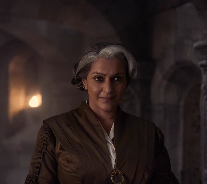 Meera Syal as Verin Mathwin stands in a dimly lit stone interior, facing forward with a calm, knowing expression. Her gray-streaked hair is neatly styled back, and she wears layered brown Aes Sedai robes with a simple pendant. Soft candlelight behind her casts warm shadows, emphasizing her composed, observant presence.
