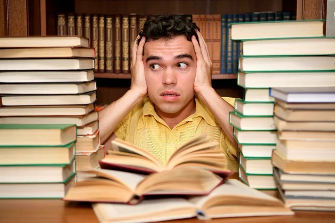 An anxious young man sitting with his hands to the side of his head among stacks of books at the library.