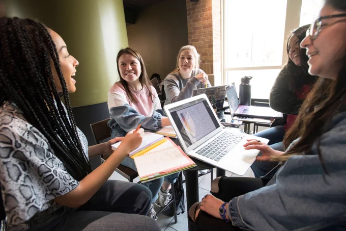 Students sitting around a table with books and laptops, talking.