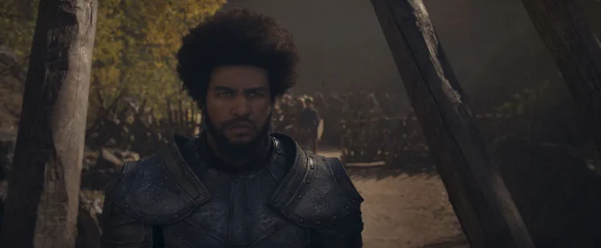Still from The Wheel of Time Season 3 showing Marcus Rutherford as Perrin Aybara standing in a forest clearing. He faces forward with a serious, focused expression, wearing dark, heavy armor that covers his chest and shoulders. His hair is worn in a full, rounded style, and his beard is neatly trimmed. Behind him, rows of armed two rivers folk stand in formation along a dirt path, partially obscured by shadows and tall wooden posts. Sunlight filters through trees in the background, creating a muted, tense atmosphere.