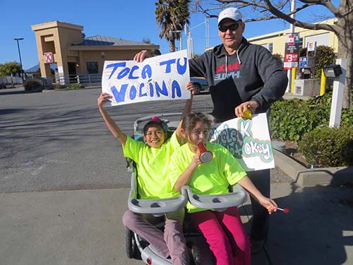 Two young kids in bright yellow t-shirts, sitting in a doublewide stroller, hold up a sign reading "TOCA TU VOCINA" ("SOUND YOUR HORN" in English), as an older man in a grey hoodie looks on smiling.