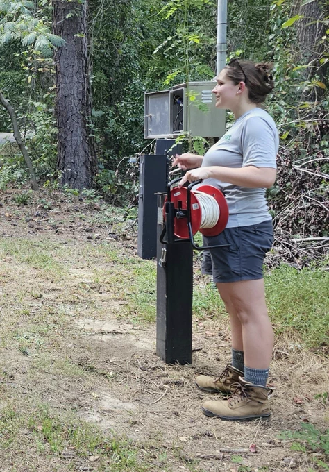 A woman holding an electric tape, standing next to a monitoring well.