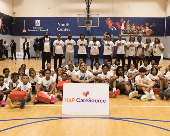Group of adults and children in a gym with basketball-themed murals, holding sports equipment and wearing matching t-shirts. A sign in the front reads "HAP CareSource."