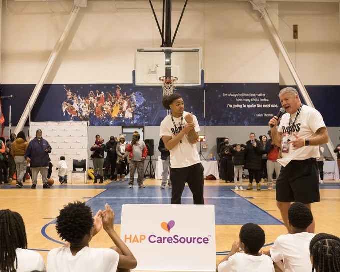 Two people, one holding a microphone, stand at the center of a basketball court during an event sponsored by HAP CareSource. A crowd of people watches in the background, with a dynamic basketball mural on the wall.