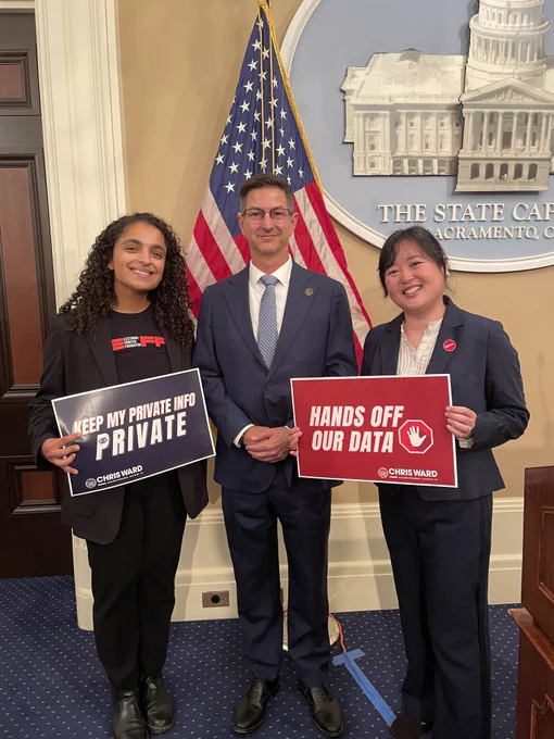 Rin Alajaji, Assemblymember Chris Ward, and Hayley Tsukayama smile in front of an American flag, holding signs reading "Keep my private info private" and "Hands off our data."