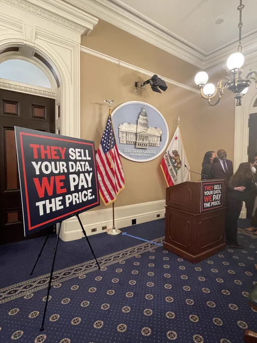 Press room with podium, US and California flags, seal of the State Capitol, and signs reading "THEY SELL YOUR DATA. WE PAY THE PRICE."