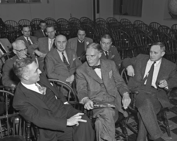Black and white image of a group of men seated in wooden chairs, engaged in conversation. Shows Governor Herter speaking with George Valley, Director Marshall Holloway sits to his left, and Jay Forrester and Robert Everett are seated near back. 