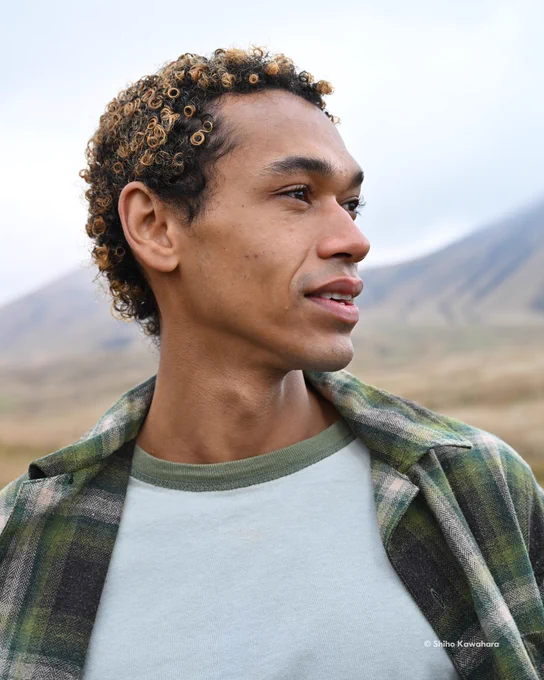 A close-up portrait of a man looking off to their right with a slight smile. He’s wearing a green plaid jacket over a white T-shirt, standing in an outdoor setting with soft-focus hills in the background. In the lower right-hand corner, on-screen text reads: “© Shiho Kawahara.” 