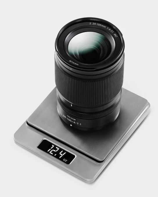 A photo of a NIKKOR Z 24-105mm f/4-7.1 lens placed upright on a silver digital scale. The scale displays a weight of 12.4 ounces. The image is shot against a clean white background. 