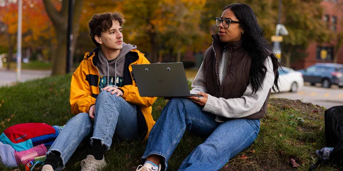 Two UWM students sit outside on campus, conversing and holding a laptop.