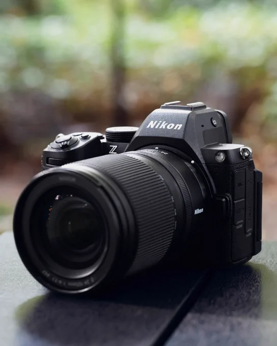  A photo of  a Nikon Z mirrorless camera with the NIKKOR Z 24-105mm f/4-7.1 lens attached, placed on a dark tabletop with soft green and brown bokeh in the background.