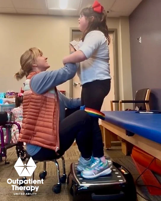 Physical therapist assists a child in standing and balance exercises using adaptive equipment during pediatric outpatient therapy.