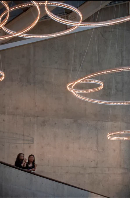 Zu sehen ist ein modernes Theater mit Wänden aus Sichtbeton mit runden, ringartigen Lichtinstallationen im gesamten Gebäude, die vom deutschen Designer Thomas Haagen als 'FLIGHT-Lichtsystem' getauft wurde. Das Lichtsystem überspannt die Eingangshalle und die Flure mit imposanten Lichtringen, die aus tausenden Soffittenlampen bestehen, die jede einzeln zwischen Aluminium-Flachprofilen mittels zweier Federn eingespannt sind. Diese Lampenhalterung für Soffittenlampen ist einmalich in der Beleuchtungswelt, die auf eine Erfindung des Lichtdesigners Thomas Haagen beruht.