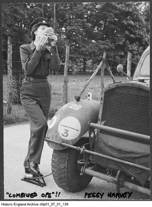 A black and white photograph of a woman in military uniform standing beside a military vehicle. The woman is applying make up and holding a compact mirror. She is pumping the tyre of the vehicle using her foot. At the bottom of the photograph is a handwritten caption which reads "Combined Ops!! Peggy Harvey".