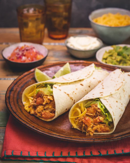 Two chicken wraps on a wooden plate accompanied by sides of guacamole, salsa, sour cream, and chips, with two glasses of iced tea in the background.