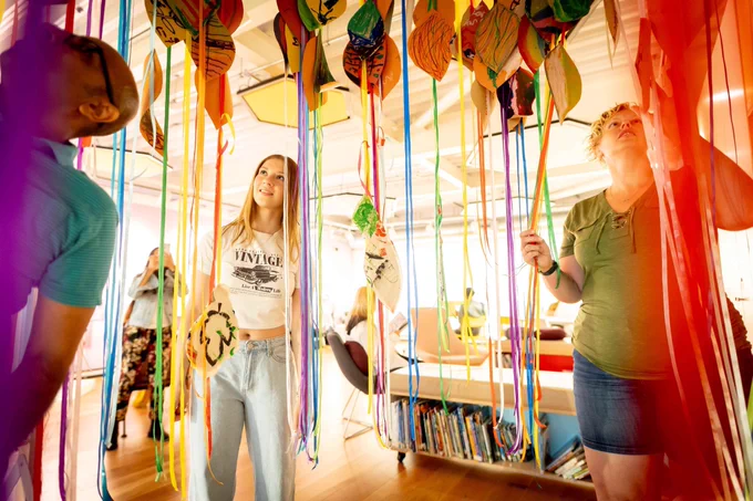 Two women and a man in a colourful art installation with colourful stripes of paper falling down around them.