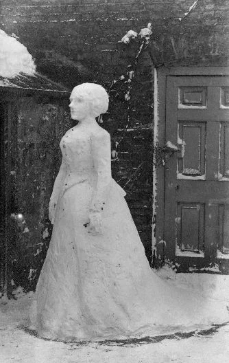 Black and white photograph taken in heavy snow, showing a life-sized snowwoman figure standing in a garden. She wears a long Edwardian-style skirt, fitted bodice and wide-brimmed hat picked out in snow, with real objects added for features. A wooden door and snow-covered trees frame the scene.