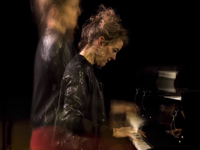 A woman with dark hair wearing a shiny dark jacket playing piano in the dark.