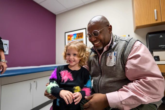 A healthcare professional wearing glasses and an ID badge smiles while interacting with a young patient in a colorful sweater in a clinic room with artwork on the walls.
