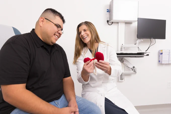 A healthcare professional in a white coat shows a kidney model to a smiling patient in an exam room.